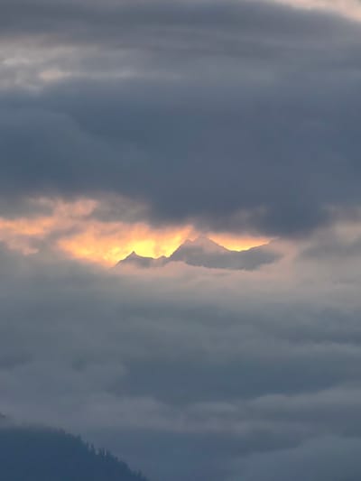 A dramatic cloudy sky frames a mountain range. The clouds are dark gray and dense, obscuring much of the view. However, a gap in the clouds reveals a bright, golden light illuminating the peaks of the mountains, creating a striking contrast between light and shadow. The mountain peaks are jagged and appear sharp against the luminous background. The lower portion of the image shows a dark, silhouetted treeline, suggesting a forest or a wooded area on the slopes of the mountains. The overall scene evokes a sense of grandeur and mystery, with the hidden mountains bathed in the glow of unseen sunlight. The location is the city of Aleo, India.