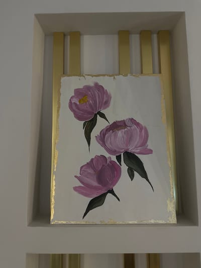 An artwork depicting three purple or pink flowers with dark green leaves on a light background is displayed within a recessed niche in a wall. The painting itself features a rough, textured gold border, suggesting gold leaf or paint applied to its edges. The flowers, possibly peonies, are rendered in a loose, painterly style. The central flower is slightly larger and more open than the two smaller ones.

The painting is supported by a series of horizontal, gold-colored metal bars or shelves. Three such bars are visible to the left of the painting, extending from the left wall of the niche to partially beneath the artwork. Similar bars are partially visible to the right, suggesting a continuous support system. The walls of the niche and the surrounding room are a plain, light neutral color, likely off-white or light gray. The overall presentation suggests an intentional and decorative display of the artwork in an indoor setting. There are no people visible, nor any discernable text within the image. The lighting appears to be artificial, casting soft shadows within the recessed area.