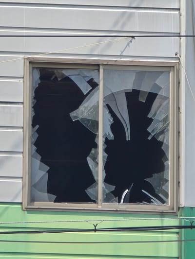 This is a close-up, eye-level exterior shot of a broken sliding window on a building. The window is divided into two panes, both of which are shattered. Large, irregular shards of glass remain attached to the frame, with significant gaps exposing the dark interior. The window is set into a wall with horizontal siding, painted a light gray. Below the window, the building has a section with bright green horizontal siding. Several wires are visible in the foreground, strung horizontally across the frame. The scene appears to be in an urban or suburban setting, possibly an industrial or commercial area. The context suggests potential vandalism or accidental damage.