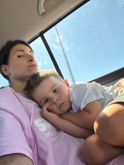 A woman with dark hair and freckles looks towards the camera with a neutral expression. A young child with light brown hair is resting their head on the woman's shoulder and chest. The child's arm is resting on their own chest, and their hand is near their face. Both are inside a vehicle. The background shows a bright blue sky visible through a window, suggesting an outdoor setting during the day. The location is identified as Kruševica, Montenegro.