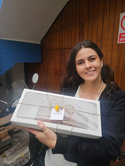 A woman is holding a rectangular white box tied with string, a yellow flower attached to the string, and a label with the text "EL PECADO DE ALE, REPOSTERÍA HORNEADA." The woman has dark hair and is wearing a dark jacket, a white top, and multiple necklaces. She is smiling at the camera. Behind her is a wooden wall with a "PROHIBIDO ESTACIONAR" sign. To the left, there is a blue wall. Part of a scooter is visible at the bottom of the image. The location is likely Magdalena del Mar, Peru, based on the label on the box.