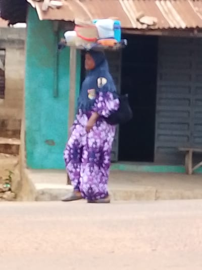 A person, wearing a hijab and a flowing purple patterned dress, balances a tray of items on their head. The person is walking on a dirt road in front of a building with a corrugated metal roof. The building is painted teal and has a dark opening that appears to be a doorway. To the right of the doorway, a wooden bench is visible. The scene is set in Iseyin, Nigeria.