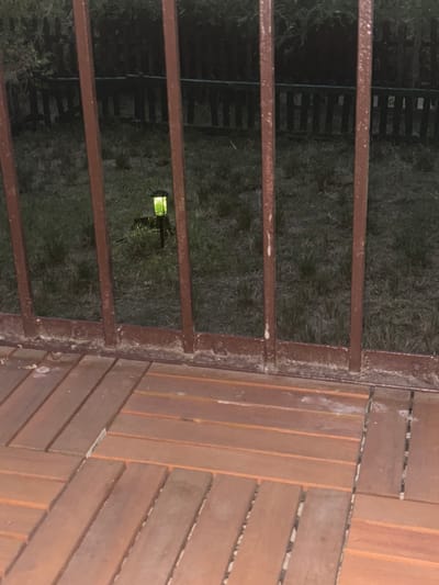 The image captures a ground-level perspective of a wooden deck with a dark metal fence in the background. A single, small solar-powered garden light with a green glow is visible on the grass beyond the fence. The deck appears to be made of wooden planks laid out in a herringbone pattern. The environment suggests an outdoor setting, possibly a backyard or patio. There are no people or animals visible in the frame. The location in Kalisz, Poland is not specified by the visual information present in the image.