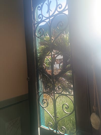 This image depicts a view through an open door with ornate wrought iron decorative elements, likely in Serravalle, Switzerland. The door opens to an outdoor scene where a large, rustic wreath made of branches is hanging. Beyond the wreath, a sunlit landscape is visible, featuring green grass and distant hills or mountains. The sun's glare is strong on the right side of the image, obscuring some details. The interior of the room shows a neutral-colored wall on the left and a dark wooden door frame on the right, with a rope and a decorative object hanging from it. There are no people present in the frame. The overall impression is one of a bright, sunny day in a natural setting, viewed from a shaded interior space.