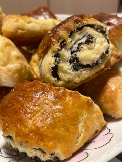 A close-up, eye-level shot showcases a pile of freshly baked poppy seed rolls, likely homemade, presented on a white plate with a delicate pink floral design. The rolls are golden brown with a slightly glossy finish, indicating they may have been brushed with an egg wash or syrup. Some rolls are whole, revealing their flaky pastry layers, while others are cut, exposing the dark poppy seed filling swirled within the dough. The texture of the pastry appears soft and yielding, with visible crumbs and a slightly uneven surface. The lighting is warm, casting soft shadows and highlighting the baked goods' appetizing appearance. The background is a blurred, neutral tone, keeping the focus squarely on the pastries. There are no people or actions depicted in the image. The exact location of Dagomys, Russia, is not discernible from the image itself.