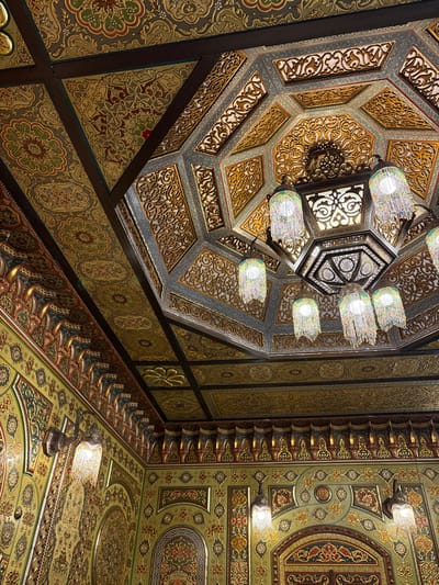 The image shows an ornately decorated ceiling featuring a central octagonal dome and adjacent flat sections. The dome is intricately carved with geometric patterns and features a hanging chandelier. The flat ceiling is decorated with floral and geometric motifs in shades of gold, green, and red. Recessed lighting is present on the flat ceiling, complementing the chandelier's illumination of the dome.