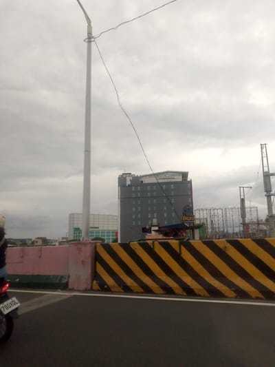The image captures an urban or semi-urban scene from a road, likely an elevated section or overpass, under an overcast sky. The overall impression is one of a cloudy day.
In the foreground, the paved road surface is visible, separated by a white line from a series of road barriers. On the left, a faded pinkish-grey concrete barrier transitions into a yellow and black striped concrete barrier that runs across the middle right of the frame. Part of a motorcycle, including its rear wheel and license plate displaying "780XQL", is visible on the far left, suggesting a vehicle in motion or parked on the road.
A tall, white street light pole stands prominently on the left side of the image, with wires attached and stretching across the sky. In the midground, beyond the barriers, a multi-story grey building with numerous rectangular windows dominates the scene. A sign on this building is visible, featuring the words "Chill" and "PADIS" (possibly "Padi's Point," a common restaurant/bar chain in the Philippines). Behind this grey building, another large, lighter-colored building can be seen. On the far right, metal framework and structures, possibly related to construction or elevated infrastructure like an MRT/LRT line, are visible against the cloudy sky.
The scene depicts a typical city environment, likely in Pasay, Philippines, given the architectural style and visible signage.
