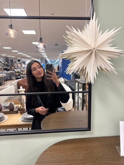 The image depicts a female subject captured in a mirror selfie within an indoor retail environment, identified as a furniture or home goods store in Pobrdje, Montenegro.
**People:** A single female, appearing to be in her 20s or 30s, is reflected from the chest up. She has long, dark hair, is wearing dark clothing, and holds a dark-colored smartphone with her right hand to take the photograph. She wears rings on her left hand and exhibits a subtle smile.
**Subjects/Objects:**
* **Mirror:** A rectangular mirror with a thin black frame occupies the central and lower right portion of the image, reflecting the scene.
* **Decorative Star:** A large, multi-pointed, cream-colored paper star ornament hangs suspended in the upper right quadrant, partially overlapping the mirror's reflection.
* **Foreground Table:** A dark wood, semi-circular table surface is visible in the lower foreground, extending from the bottom edge of the mirror.
* **Reflected Store Interior:** The mirror reflects a broad view of a retail space containing various furniture displays. Visible items include multiple sofas and armchairs in neutral tones (cream, grey, black), several tables, and shelving units.
* **Reflected Table Setting:** To the left of the female's reflection, a wooden table is set with placemats, grey bowls, and dark, textured decorative objects resembling pinecones.
* **Lighting:** Two clear glass pendant lights with exposed filaments hang from the ceiling, along with recessed fluorescent lighting fixtures. A red fire alarm or sensor is visible on the distant ceiling.
**Actions:** The female is actively taking a self-portrait using her smartphone, positioning the device to capture her reflection in the mirror.
**Environment:** The scene is well-lit, suggesting artificial illumination characteristic of a commercial showroom. The background features structured displays of home furnishings against light-colored walls and flooring, indicating an organized retail setting designed for product presentation.