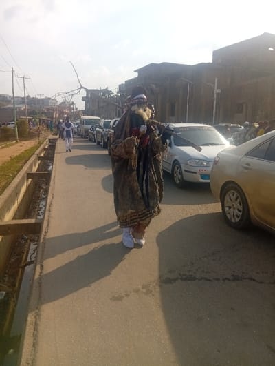 The image depicts a daytime street scene at Itakpe Junction, Nigeria. In the foreground, slightly left of center, a single individual stands or walks, adorned in an elaborate, multi-layered dark costume. This costume features a wide-brimmed dark hat with white and red fabric accents, a masked face with a white beard-like element, and a long, dark, rough-textured garment reaching to the ankles, covering white shoes. The figure holds a white cup-like object in the left hand and carries a slender, dark stick horizontally across the right shoulder. A distinct shadow is cast to the figure's right on the asphalt.

To the left of the main figure, a concrete drainage ditch runs parallel to the paved road, containing some debris. Beyond the ditch, a dirt shoulder and utility poles with overhead lines are visible. Further down the road on the left, one person dressed in white clothing walks away from the camera, and other indistinct figures are present in the distant background.

On the right side of the paved road, a line of vehicles is stationary or moving slowly. The closest visible vehicle is a silver sedan with a sticker on its windshield, followed by a partially visible gold sedan. A white van and other cars extend into the background. Buildings of varying styles, including a prominent large, dark, multi-columned structure, are visible in the far background. The sky above is clear and light, indicating bright daylight.