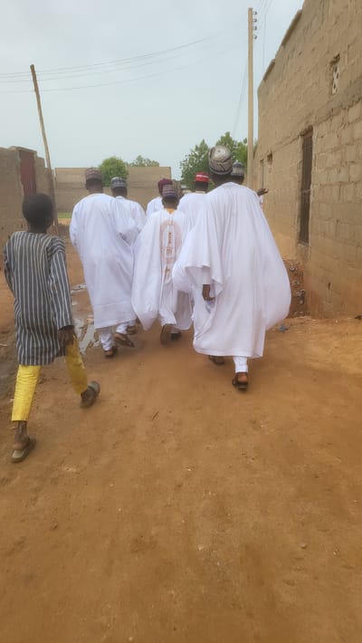 Seven males walk along an unpaved reddish-brown dirt path in Jogana, Nigeria, under an overcast sky. Six adults, viewed from behind, wear white, flowing traditional garments and various patterned kufi-style hats. One adult's garment features a brown embroidered design on the back. They are walking away from the camera. A young boy walks ahead of and to the left of the adult group, also viewed from behind, wearing a striped long-sleeved shirt and yellow trousers, with sandals on his feet. The path shows some scattered debris and a small wet patch to the left. To the left of the path, a light-colored wooden utility pole rises vertically. To the right, a row of light-brown, unrendered brick or mud-brick buildings with visible windows and doors extends into the distance. Utility wires span horizontally across the upper portion of the frame. Green foliage from trees is visible behind the buildings in the middle distance.
