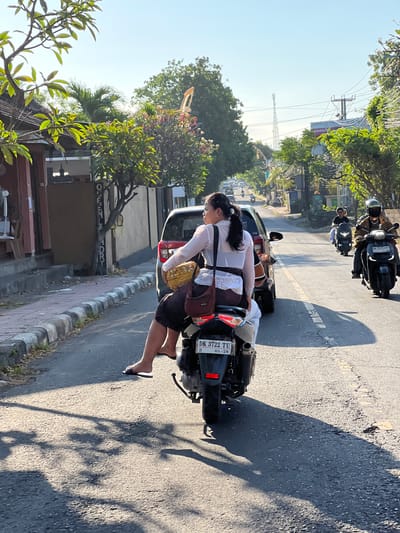 In the city of Kubu, Indonesia, a woman is riding on the back of a scooter. She is wearing traditional Balinese clothing, including a white blouse and a dark skirt. A woven basket is slung over her shoulder. She is sitting sideways on the scooter, with her legs crossed and her feet resting on the side. The scooter has a license plate that reads "DK 3722 TU." The scooter is in motion on a paved road, which is lined with trees and buildings. Other scooters and a car are visible in the background, indicating traffic. The sun is shining, casting shadows on the road. The overall scene depicts a typical day in a bustling Indonesian city.