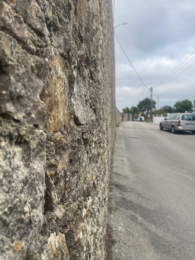 The image shows a close-up of a rough, grey stone wall in the foreground, with a paved road and a silver car in the background. The road recedes into the distance, with houses and trees lining its sides. A street lamp and power lines are also visible. The sky is overcast. The location is identified as São João das Lampas, Portugal.