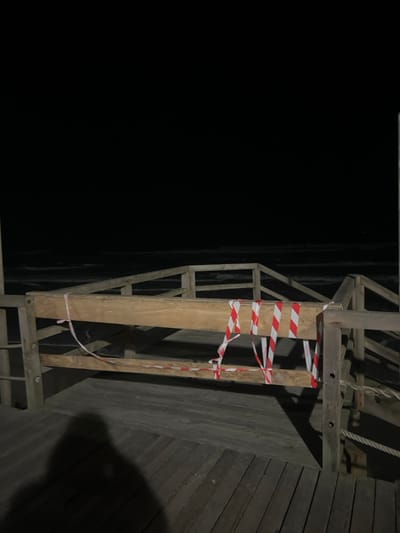 A dimly lit, angled shot captures a wooden structure, possibly a ladder or scaffolding, secured with red and white striped warning tape. The structure is attached to a wall made of horizontal wooden planks. To the left, a large expanse of darkness suggests an open space or drop-off. The composition is stark, with the wooden structure and the tape standing out against the dark background.
