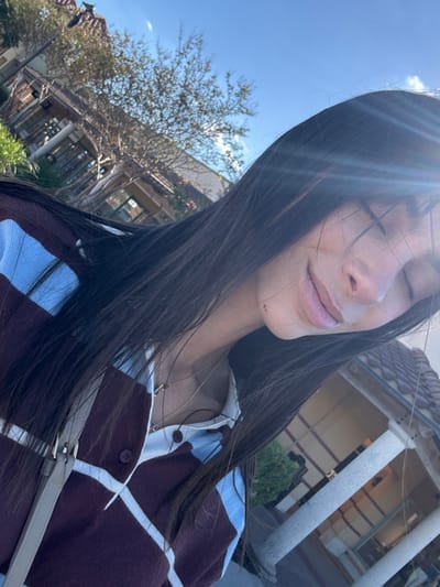 A woman with long, dark hair is facing the sun with her eyes closed. She wears a burgundy and light blue striped cardigan and has two necklaces. In the background, there is a building with columns, trees, and a blue sky. The sunlight casts a bright glare on the image.