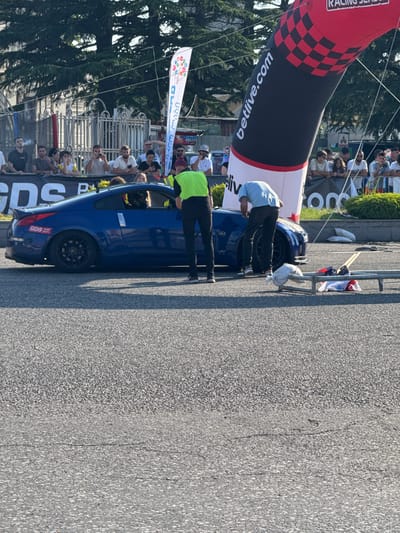 In Poti, Georgia, a blue Nissan 350Z is parked on an asphalt road, likely near a race track or event venue, as indicated by the inflatable archway with "betlive.com" branding and checkered pattern in the background. Spectators are visible behind a barrier in the upper portion of the image. Two individuals are attending to the car. One person, wearing a bright green vest, stands by the open driver's side door. Another individual, in a light blue shirt, is bent over near the front passenger side of the car. A metal frame with sandbags and flags is positioned on the ground to the right of the vehicle. The environment suggests an outdoor event with a focus on motorsports.