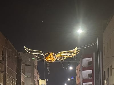 The image shows an outdoor scene at night in the city of Berrechid, Morocco. The primary subject is a decorative light display suspended across a street, featuring a crown within a circular frame, flanked by illuminated wings. The lights are golden yellow and white, with hanging strands of smaller lights. The display is strung between buildings on either side of the street. The buildings appear to be multi-story structures, some with visible windows and balconies. Streetlights are also present, casting a glow. The sky is dark, indicating it is nighttime. The overall scene suggests a festive or decorative illumination for a street or event.