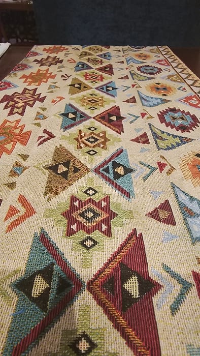A tapestry featuring geometric, Southwestern-inspired designs is prominently displayed. The patterns consist of various triangles, diamonds, and starbursts in hues of red, blue, green, brown, and orange. These motifs are arranged in rows, creating a repeating design on a beige woven background, showcasing the fabric's texture and intricate detailing. The tapestry's side is visible on the left, near a dark blue wall.