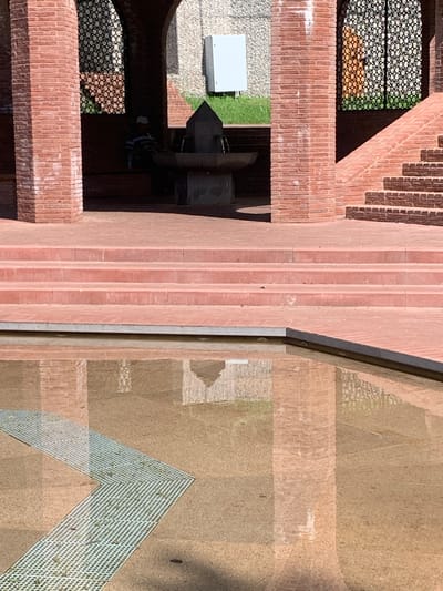 The image shows a reflective pool in front of a brick structure with arched openings and a set of stairs. A person wearing a striped shirt and a hat is seated inside one of the arched openings. In the pool, the reflection of the brick structure, stairs, and the sky is visible. There is a metal grate in the foreground of the pool, forming a diagonal line. The scene appears to be an outdoor public space. The specific location in Arbuzov-Baran, Russia cannot be determined from the image alone.