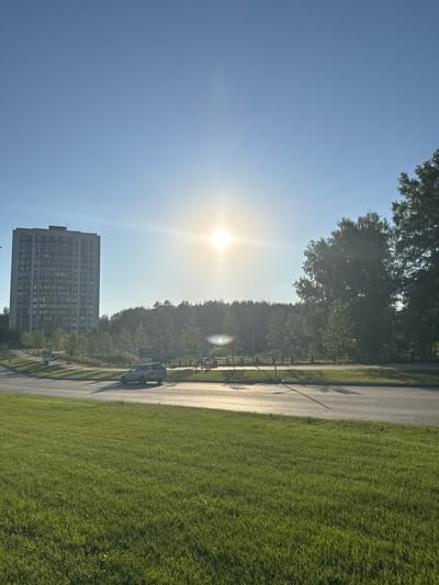 This media file depicts an outdoor scene featuring a tall, grey apartment building with numerous windows, a paved pathway, and a lush green lawn. The sun is visible and bright, casting lens flare across the image, suggesting it is either early morning or late afternoon. Trees line the pathway and are also present behind the apartment building. A white van is parked on the side of the pathway, and a fence runs alongside it. The sky is clear and blue. The orientation of the image is such that the apartment building appears to be in the lower-left quadrant, with the pathway and lawn stretching to the right. The overall impression is a tranquil, sunlit day in a suburban or park-like setting. No text is visible in the image.