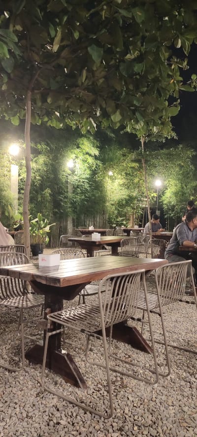 This image depicts an outdoor dining area at night, likely a restaurant or cafe, in Bireuenmeunasahdayah, Indonesia. The scene is illuminated by artificial lighting, casting a warm glow on the gravel ground, wooden tables, and metal chairs. Lush green bamboo plants form a dense backdrop, creating a natural and slightly enclosed ambiance.
In the foreground, a wooden table with a white tissue box sits prominently, surrounded by several empty metal chairs. The table has a slatted wooden surface and sturdy wooden legs. Beyond this table, other similar tables and chairs are arranged, some occupied by patrons.
In the mid-ground, a few individuals are visible seated at tables. One person on the right is wearing a grey shirt and appears to be looking down at their phone or a menu. Another individual, also in the mid-ground and on the right side, is wearing a dark shirt and is seated, seemingly engaged with something on the table. Further back, more tables and chairs are set up, with some people visible in silhouette.
The environment is characterized by the presence of trees with large green leaves overhead, their branches providing a canopy. The ground is covered with light-colored gravel. The background is dominated by the tall, slender stalks of bamboo, which are illuminated by spotlights, creating a vibrant green contrast. Multiple light sources are visible, including globe-shaped lamps hanging from trees and possibly wall-mounted lights. The overall impression is one of a relaxed, casual dining setting, designed to be an outdoor escape.