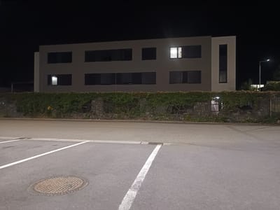 A nighttime exterior scene in Milovice, Czechia, shows a modern, multi-story building with a flat roof and a light-colored vertical section on the right. The building facade is dark gray with rows of windows, some of which are illuminated from within, displaying horizontal blinds. A stone wall topped with dense green ivy runs along the front of the building, separating it from a paved area in the foreground. This paved area includes parking lines and a manhole cover. A street lamp is visible to the right of the building, casting light on the surroundings. The sky is dark and clear.