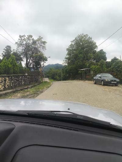 This image depicts a road in Skurdi, Georgia. The scene is viewed from inside a vehicle, looking out through the windshield and dashboard. A dark-colored sedan is parked on the right side of the road, facing away from the camera, with a license plate visible. On the left side of the road, there is a stone wall bordering a property with manicured coniferous trees. Further down the road, a paved surface transitions to a dirt or gravel area. Tall trees line both sides of the road, and a lamppost is visible. In the background, a hillside covered in vegetation suggests a rural or semi-rural setting. The sky is overcast and cloudy.