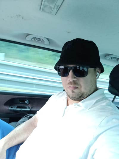 The image shows a man sitting in the backseat of a car, wearing a black bucket hat, sunglasses, and a white polo shirt. He has a light beard and is wearing a white wireless earbud in his right ear. The man is looking directly at the camera, with a neutral expression.
The setting appears to be a car interior, with the car's ceiling and window visible. The window shows a blurred view of green foliage and what appears to be a light blue sky, suggesting it is daytime and possibly sunny. The car's interior includes a grab handle and a door panel. The lighting is bright, indicating it is daytime.
The man's posture and attire suggest he is traveling. The reflection in his sunglasses shows a person taking the photo, also in the car. The overall mood is casual and relaxed. No text is visible in the image. The location context provided is Ostředek, Czechia, which aligns with the appearance of a car interior.
