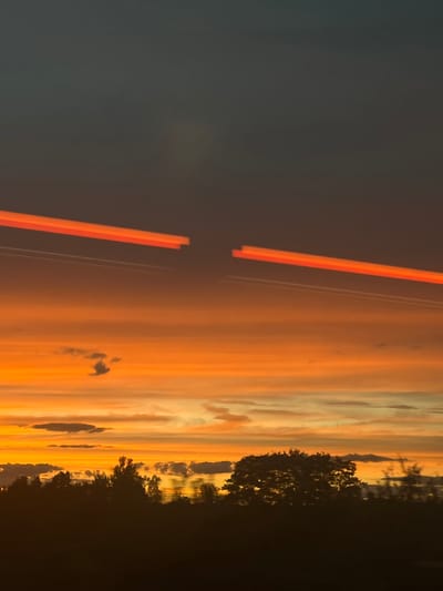 The image depicts a sunset over Baiersdorf, Germany. The sky is a gradient of dark blue at the top transitioning to vibrant oranges and yellows closer to the horizon. Two distinct orange light streaks are visible in the upper portion of the sky, suggesting the passage of vehicles or aircraft. Below the sky, a silhouetted treeline runs across the bottom of the frame, with some darker clouds interspersed within the sunset colors. The overall impression is one of a serene natural scene, possibly viewed from a moving vehicle given the light streaks.