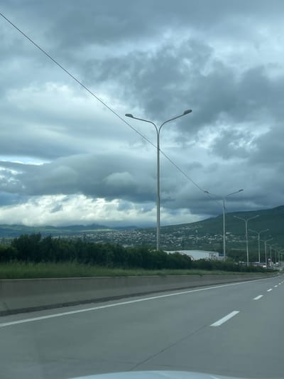 The image depicts a view from a vehicle traveling on a multi-lane highway. Adjacent to the highway is a line of dense, green trees. Beyond the trees is a city, possibly Gorovani, Georgia, situated at the base of a large, green mountain. Above, a cloudy, overcast sky dominates the view. Streetlights line the highway. White lane markings are visible on the pavement.