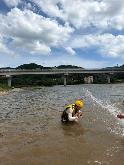 In the city of Seo-myeon, South Korea, a person wearing a yellow helmet and a yellow life vest is kneeling in a muddy river, splashing water. The person is wearing a white long-sleeved shirt under the life vest. The river is wide and appears to be shallow near the bank. In the background, a bridge spans across the river, supported by concrete pillars. Trees and hills covered with vegetation are visible beyond the bridge. The sky is partly cloudy with white clouds against a blue backdrop. The overall scene depicts an outdoor recreational activity on a warm day.