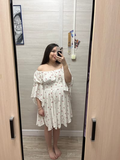 The scene takes place indoors, within a private residence in the city of Kozlovka, Russia. A young adult female is depicted in a full-length mirror selfie. She has long, dark hair, extending past her shoulders, and exhibits a faint smile. She is wearing a white, off-the-shoulder short dress adorned with a repetitive pattern of small red cherries. The dress features wide, ruffled sleeves and a defined waistline. A thin, light-colored necklace is visible around her neck, and a simple bracelet is on her left wrist. She stands barefoot on an unseen floor. Her right hand holds a light-colored smartphone, positioned to capture her reflection, with the device partially obscuring the lower right portion of her face. The mirror is framed by light-colored wooden panels, likely part of a wardrobe or closet, each fitted with a dark, rectangular handle. The wall visible in the reflection behind the subject is light-colored and horizontally paneled. To the left of the subject's reflection, a dark-framed, vertical artwork depicting a human face is mounted on the wall. To the right, a collection of small objects, including what appears to be a ruler or thermometer, several decorative crystal strands, and small photographs or cards, are affixed to the wall.