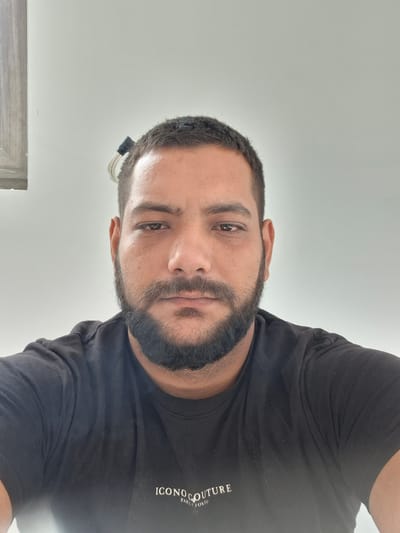 The image is a close-up, head-and-shoulders selfie of a man with dark hair and a full beard. He is wearing a black t-shirt with a white logo that reads "ICONOCULTURE PARIS TOKYO." The background is a plain, light-colored wall. There is a wooden frame on the left side of the image, suggesting the presence of a window or a piece of furniture. A small, black object with white wires is visible in the man's hair on the right side of his head. The man's expression is neutral. The image does not contain any specific visual cues to place it in Dolna Malina, Bulgaria; the description provided in the prompt is not reflected in the image.