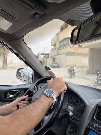 The image captures a first-person perspective from within a vehicle, looking forward through the windshield in Harasta, Syria. The driver's forearms and hands are positioned on a black steering wheel; a white smartwatch with a gold bezel is worn on the right wrist, and a ring is visible on the right ring finger. The vehicle's interior visible includes a grey dashboard, a chrome door handle on the left door, and a rearview mirror mounted on the windshield.

The exterior scene depicts an unpaved, dusty road surface. Buildings line the street, some displaying signs of damage or construction, including a structure on the left with exposed framing and a multi-story building to the right featuring blue-tinted windows on a second-floor balcony. Further down the road, a dark-colored sedan is parked, facing away from the viewer. Two motorcycles are present on the right side of the street; one is a red and black motorcycle, and a second, lighter-colored motorcycle is partially obscured to its right. An extensive light-colored fabric canopy or tarp structure is suspended overhead across the street. The sky appears hazy or overcast. Sparse, dry vegetation is visible on the far left.