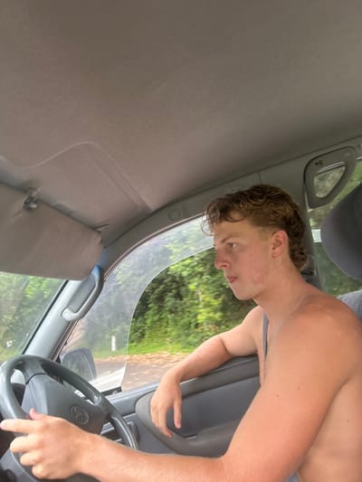 Inside a car, a shirtless, tan-skinned male with curly hair, is seated in the driver's seat. His eyes look towards the window with a serious expression. The car's interior is predominantly light gray. A glimpse of green foliage is visible through the car's window, suggesting an outdoor setting. The steering wheel is in his grasp, indicating he is driving.
