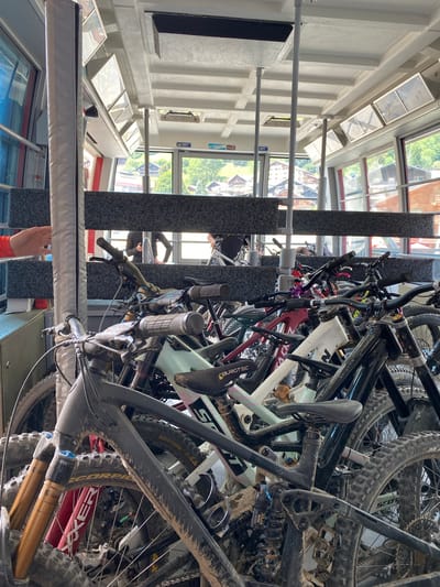 The image depicts a rack overflowing with mountain bikes inside a building, likely a cable car station, as suggested by the interior view of Champéry, Switzerland, visible through the windows. The bikes are piled haphazardly, showing signs of heavy use with mud and dirt covering their frames, wheels, and components. The color palette is predominantly dark, dominated by the black and gray of the bicycles. The environment is a mixture of industrial and outdoor elements, with the interior of the cable car station juxtaposed against the green landscape outside. Evidence of human activity is apparent, with people reflected in the windows in the background, although their actions cannot be explicitly determined.