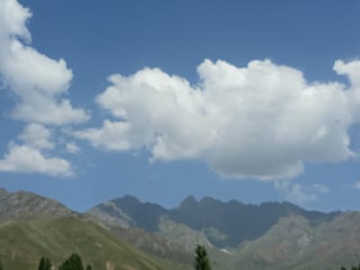 The image depicts a wide landscape under a bright blue sky with scattered white clouds. In the foreground, lush green rolling hills lead up to a range of jagged, rocky mountains in the background. Several trees are visible at the base of the hills. The scene is tranquil and suggests a rural or mountainous environment. The provided text states that the location is the city of Kalaynay, Tajikistan, which would place this scene within that geographical context. There are no people or specific actions depicted in the image; it is a static landscape view.