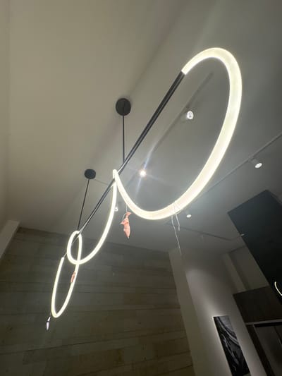 A modern ceiling light fixture with three intersecting illuminated rings is mounted on a white ceiling. The light fixture is supported by thin, dark metal rods connected to circular mounting plates. Small, pink or reddish objects are near two of the rings. The adjacent wall has a textured, stone-like appearance. Other ceiling fixtures and decorative elements are visible in the background. The location and city are not apparent from the image.