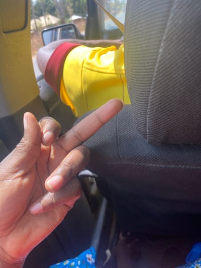 The scene is captured inside a vehicle in Unguwar Gaya, Nigeria. In the foreground, a dark-skinned left hand is raised, forming a "V" gesture with its extended index and middle fingers, while the thumb, ring, and pinky fingers are bent. The hand shows skin texture and highlights from ambient light. To the right, a dark gray, ribbed fabric car seat headrest occupies the midground. Behind and to the left of the headrest, a person is seated in the front passenger or driver's seat, wearing a bright yellow short-sleeved garment and red shorts or an undergarment with a red waistband. Their left arm is visible, bent at the elbow, with the forearm resting on their leg. Through the vehicle's window, the background reveals an outdoor environment consisting of a dirt road and sparse green vegetation under bright, natural daylight.