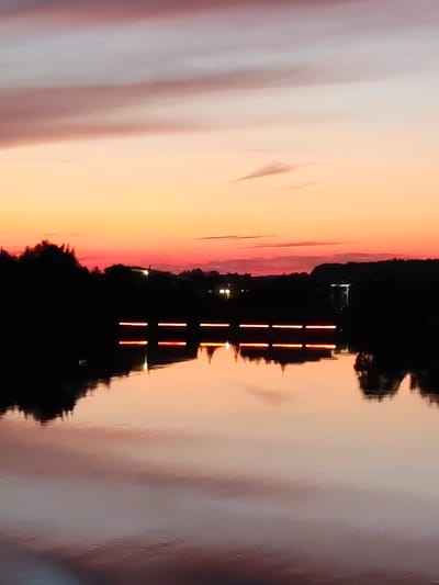 A serene sunset over a body of water near Konakovo, Russia, features a fiery orange and pink sky reflected in the calm surface. Silhouetted trees and distant lights form a dark line along the horizon. Several bright, horizontal red lights are visible just above the water's surface, creating a reflection below. The overall scene evokes a peaceful and tranquil atmosphere.