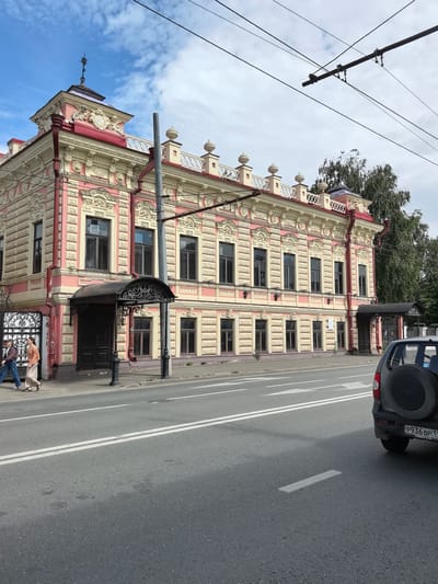 This media file captures a street view in Usady, Russia, under a partly cloudy sky. The scene features an ornate, multi-story building with a pink and cream facade, characterized by detailed architectural embellishments, including decorative window frames, cornices, and a small turret with a spire. The building appears to be a historic structure, possibly a mansion or government building, with multiple windows and several entrances.
In the foreground, a paved road with white lane markings runs horizontally across the frame. A dark-colored SUV with a spare tire mounted on the back is visible on the right side of the road, partially obscuring the building's right side. On the sidewalk in front of the building, two individuals are walking from left to right. One is wearing a light-colored outfit, and the other is in darker clothing. Overhead, electrical wires for what appear to be a tram or trolley system crisscross the sky, suggesting a public transportation route.
Notable details include the architectural style of the building, the presence of overhead wires, and the general appearance of a town or city street. The lighting suggests it is daytime, with a mix of sun and clouds. No specific events are depicted, but the scene portrays a typical day on a street with historical architecture and active traffic. There is visible text on the license plate of the SUV, which reads "P936 BP 11".