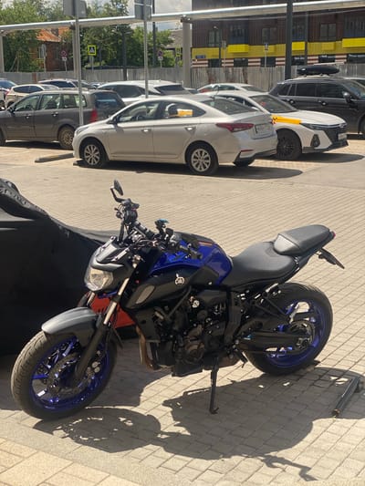A blue and black motorcycle is parked on a paved area next to several cars. The motorcycle is in the foreground, with its front wheel facing right. It has a sporty design with exposed engine parts and a blue rim on the rear wheel. A black motorcycle cover is partially visible in front of the motorcycle.
To the left of the motorcycle are several cars parked in a row. The closest car is white, with its rear end facing left. Further down the line are a dark gray car and other vehicles. The cars are parked on a paved surface that appears to be a street or parking lot.
In the background, there is a building with yellow and brown colored panels. Metal pipes run along the side of the building. Trees and some signage are also visible.
The scene is illuminated by sunlight, casting shadows on the pavement. The time of day appears to be daytime, likely morning or afternoon, given the strong shadows. The weather seems pleasant, with clear skies implied by the bright sunlight. The location context is Kotelniki, Russia. No people are visible in the image. There is some text visible on the license plates of the cars, but it is not legible. A logo, possibly of a manufacturer, is visible on the side of the motorcycle.