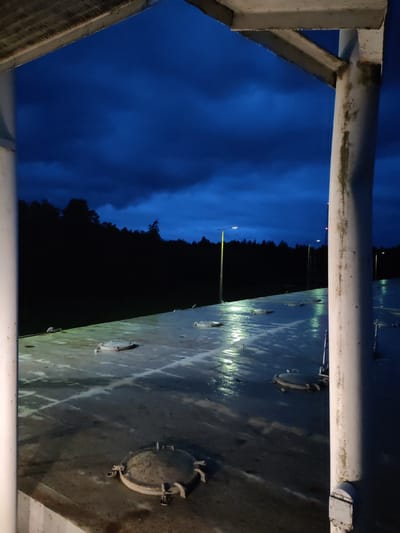The image shows a desolate, industrial landscape at dusk or early morning. The sky is a deep, ominous blue with dark clouds. A forest line is visible in the distance, silhouetted against the dim light. In the foreground, a wet, flat surface, possibly a road or platform, is covered with several circular hatches or manhole covers, some of which are rusty. The surface is reflective, suggesting it may have recently rained or is wet from dew. Two large, white, cylindrical support beams frame the scene, one on the left and one on the right. A metal structure, possibly part of a roof or awning, is visible at the top left of the frame. Streetlights are visible in the distance, illuminating the dark environment. The overall atmosphere is one of stillness and isolation. The location is identified as Devyatiny, Russia. No people or actions are visible in the scene.