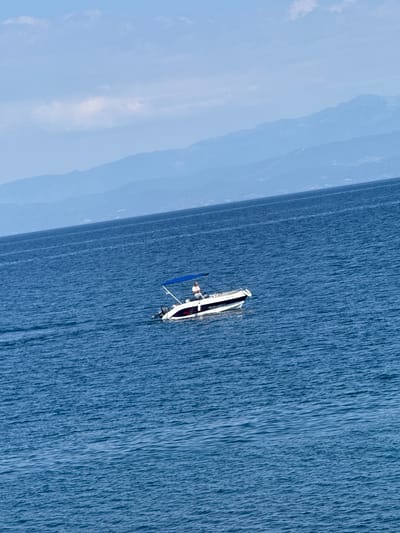 A man stands on a white speedboat with a blue canopy navigating the deep blue sea. The boat is positioned in the middle of the frame, with a slight tilt to the left. In the background, a hazy outline of mountains stretches across the horizon under a light blue sky with scattered white clouds. The water is slightly rippled, indicating a calm day. This scene likely depicts a leisure activity near Skala Marion, Greece, given the coastal mountainous landscape.