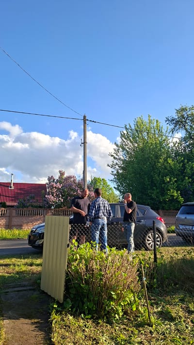 The image depicts an outdoor scene in Сваткаўскі сельскі Савет, Belarus, under a bright blue sky with scattered white clouds. Three men are standing near a chain-link fence, seemingly engaged in conversation or observation. One man is wearing a black t-shirt, another a blue and white plaid shirt, and the third also wears a black t-shirt. Behind them, a dark-colored vehicle is partially visible. To the right of the men, on the other side of the fence, is a bush with green and brown foliage. In the background, a red-tiled roof and a brick wall can be seen, along with a lilac bush and trees. An electric pole with wires extending across the sky is also visible in the foreground. A sheet of corrugated metal is leaning against the fence. Grass and other foliage line the ground.