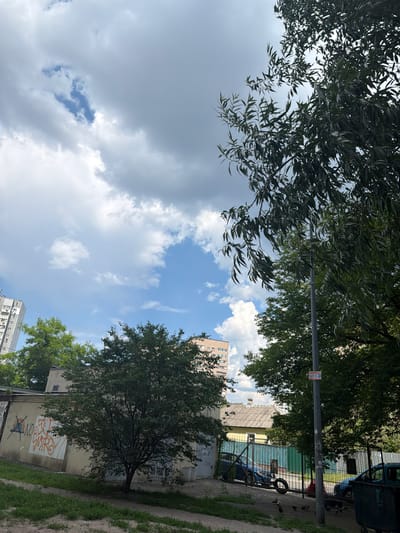 This media file captures an outdoor scene, likely in Kyiv, Ukraine, during daytime. The perspective is oriented sideways, looking upwards. Lush green trees with dense foliage dominate the foreground, framing a sky filled with a mix of dramatic gray storm clouds and bright blue patches with fluffy white cumulus clouds.
In the mid-ground and background, glimpses of urban infrastructure are visible. A multi-story apartment building with a brick facade and numerous windows stands to the right, its light-colored exterior contrasting with the greenery. Adjacent to it is a weathered building with a yellow roof, marked with graffiti. A tall lamppost with a sign attached to it is also present. Further into the background, there is a glimpse of what appears to be a fence and possibly some vehicles, including a large dark vehicle with a weathered appearance. A discarded tire is visible near the fence.
The overall atmosphere suggests a typical urban environment, possibly a residential area with surrounding trees. The presence of both dark clouds and blue sky hints at changeable weather, possibly a break in a storm or the approach of one. The graffiti on the buildings suggests a lived-in, possibly older part of the city. No specific activity or event is clearly depicted; the scene appears to be a still moment of an outdoor setting. The image does not convey any strong emotions, presenting a straightforward observation of the environment.
The sign on the lamppost, though partially obscured and sideways, contains visible Cyrillic text, typical for Ukraine. While not fully legible, the characters suggest a public announcement or advertisement.