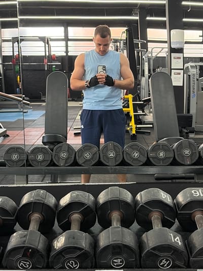 A man is standing in a gym, taking a selfie in a mirror. He is wearing a light blue sleeveless shirt, dark blue shorts with a white Nike logo, and black weightlifting gloves. The gym appears to be a commercial fitness facility with various weight machines and equipment visible in the background. In the foreground, a rack of dumbbells is prominently displayed, with weights ranging from 10 to 16. The man is holding a smartphone in his hands, with the screen facing towards himself. The lighting in the gym is bright, likely from overhead fluorescent lights. There is no indication of the specific location within Lelystad, Netherlands, other than it being a gym.