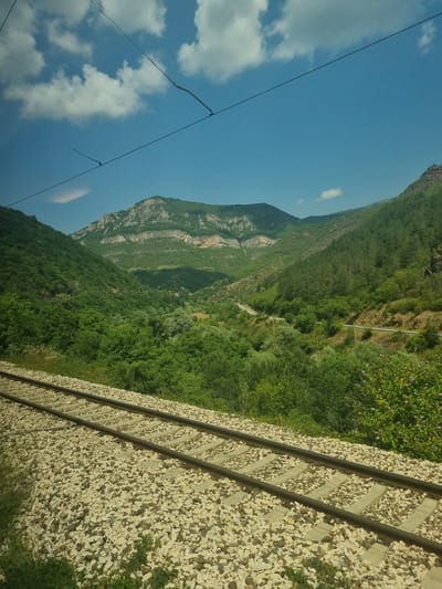 The image depicts a scenic view of a mountain valley with a train track in the foreground. The train track, composed of dark rails and light-colored ballast, runs diagonally across the lower portion of the frame. Beyond the tracks, the landscape is dominated by lush green trees and vegetation, indicating a rural or natural setting. In the mid-ground, a road winds its way through the valley, bordered by trees on either side. The background features a majestic mountain range with layered rock formations and vegetation. The sky above is a clear blue, dotted with scattered white clouds. The overall impression is one of natural beauty and tranquility. The provided location of Ochindol, Bulgaria, suggests this is a view from or near that area. There are no people or animals visible in the image. The scene is captured in daylight, with clear visibility and natural lighting.