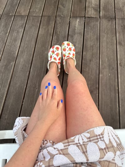The image shows a person's legs and feet, adorned with strawberry-patterned Crocs. The person's fingernails are painted a bright blue. They are seated on a white lounge chair, draped with a beige and white checkered towel, on a wooden deck. The background consists of wooden planks. The scene is set outdoors, possibly in Yuzhnyye Koryaki, Russia.