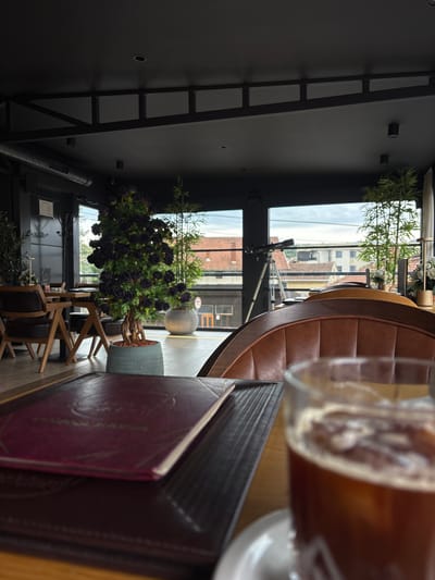 The image depicts the interior of a modern cafe in Loznica, Serbia. The cafe features dark ceilings with exposed industrial elements, including ventilation systems and lighting fixtures. The walls are also dark, creating a contemporary aesthetic. In the foreground, a table is set with a dark brown placemat and a burgundy menu. To the right, a glass of iced coffee is prominently displayed, with ice cubes and a foamy head visible.
The middle ground of the image reveals a seating area with comfortable-looking brown leather chairs. A large potted plant with dark purple roses stands near the window, adding a touch of color to the scene. Behind it, a tall bamboo-like plant is also visible. The cafe has large windows that offer a view of the city's rooftops and buildings, suggesting an urban setting. A telescope on a tripod is positioned near the window, hinting at a potential interest in astronomy or a unique vantage point from the cafe.
The background shows a glimpse of the city outside, with terracotta roofs and various buildings. The sky appears overcast. The overall ambiance suggests a relaxed and stylish cafe, possibly a place for patrons to enjoy drinks, socialize, or work.