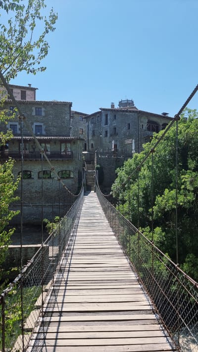 The image shows a pedestrian suspension bridge made of wooden planks and metal mesh railings. The bridge spans across a gorge or river, leading towards a cluster of stone buildings that appear to be historic. The architecture suggests a European setting, specifically the medieval village of Rupit i Pruit, Spain, known for its ancient stone houses and narrow streets. Lush green trees surround the bridge and buildings, indicating a natural environment. The sky is clear and blue, suggesting a sunny day. There are no people visible in the image. The perspective is from the beginning of the bridge, looking towards the village.